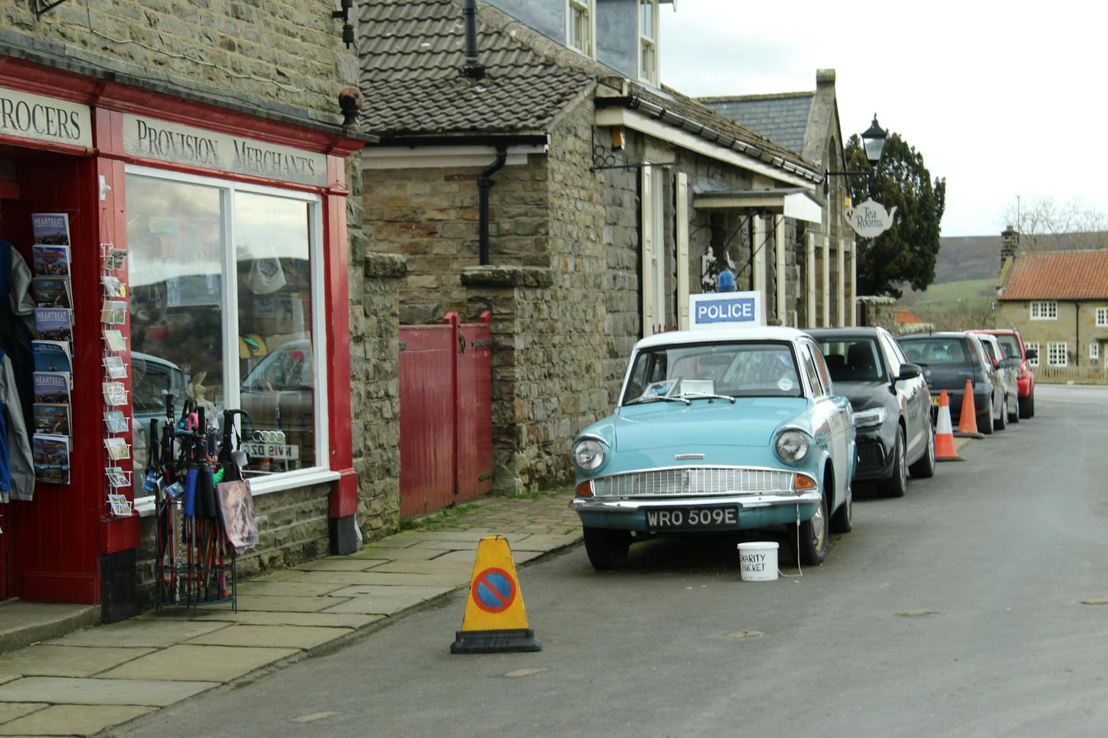 A pale blue Ford Anglia 105E in period police livery parked on a stone-built English village street