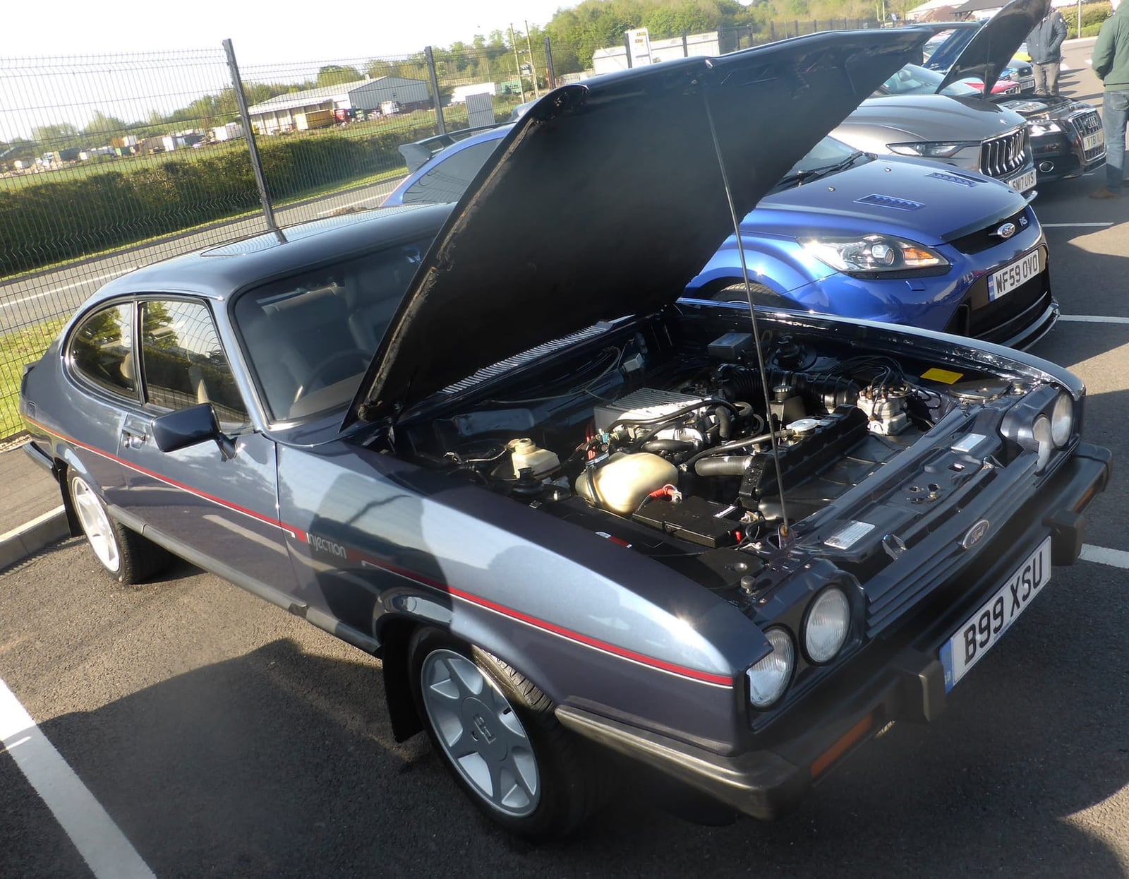 A grey 1984 Ford Capri 2.8 Injection Special with its bonnet raised at a show, the fuel-injected Cologne V6 visible