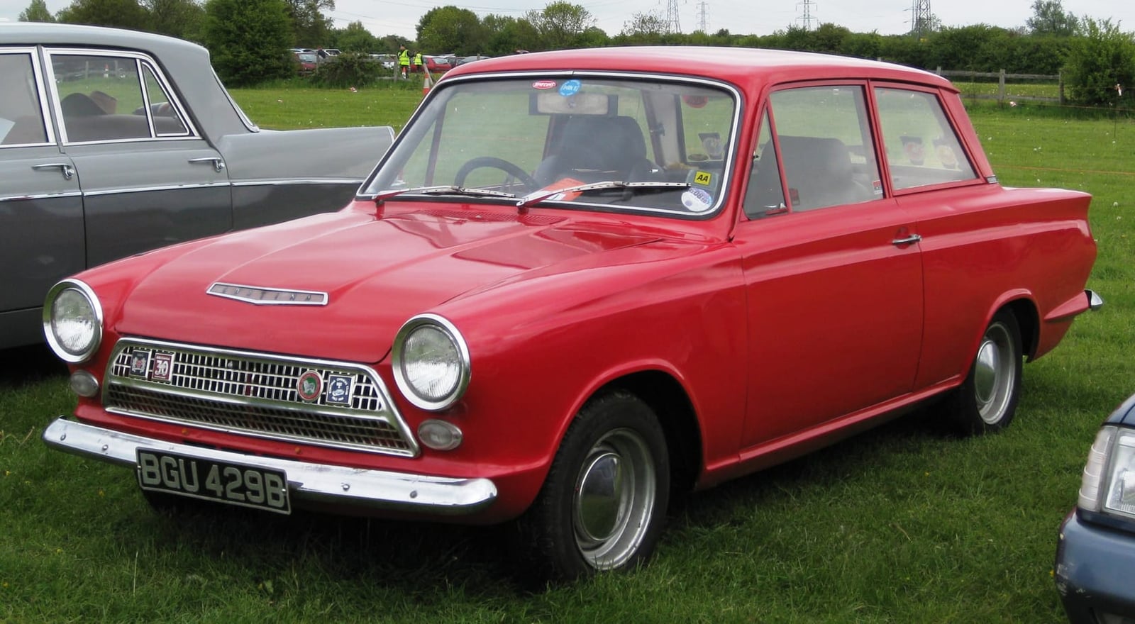 A red 1964 Ford Cortina Mk1 two-door on grass at a classic car show, front three-quarter view, UK registration SGU 429G