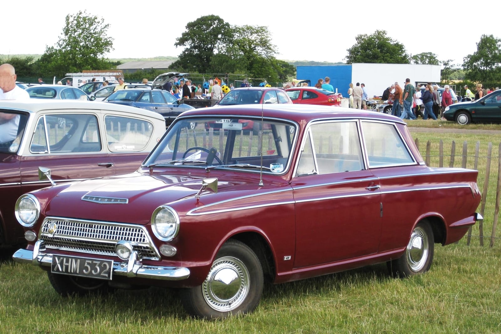 A maroon pre-facelift Ford Cortina Mk1 two-door on grass at a classic car show, front three-quarter view, UK registration XHM 332