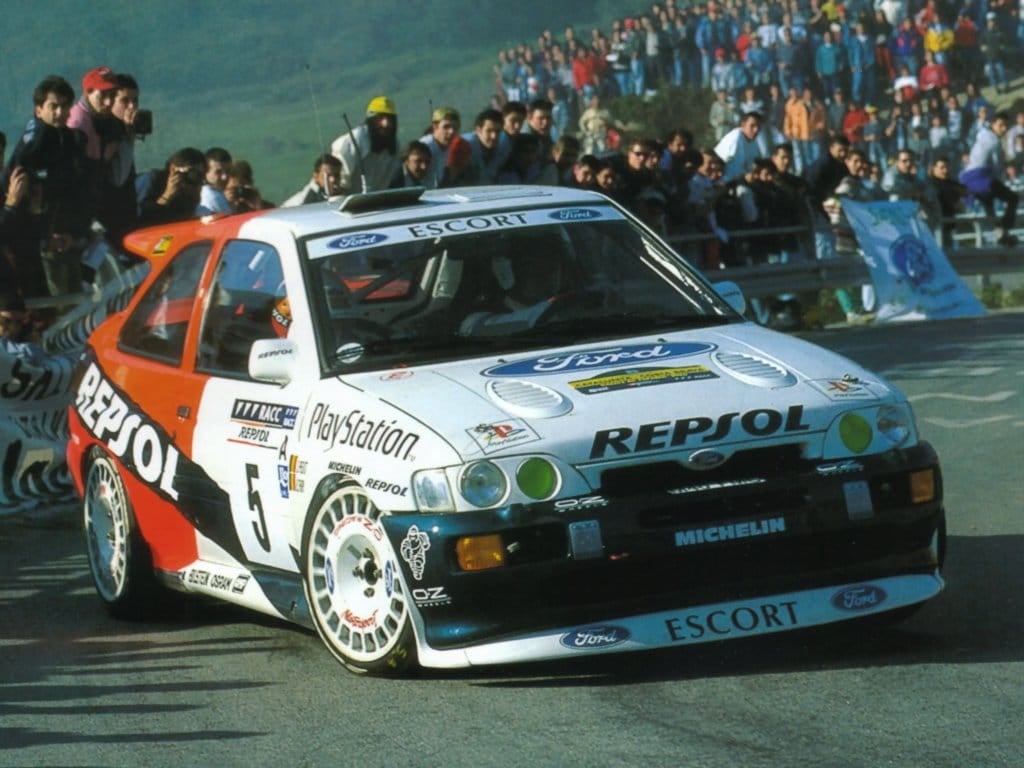 A Repsol-liveried Ford Escort RS Cosworth rally car on a special stage with spectators lining the road