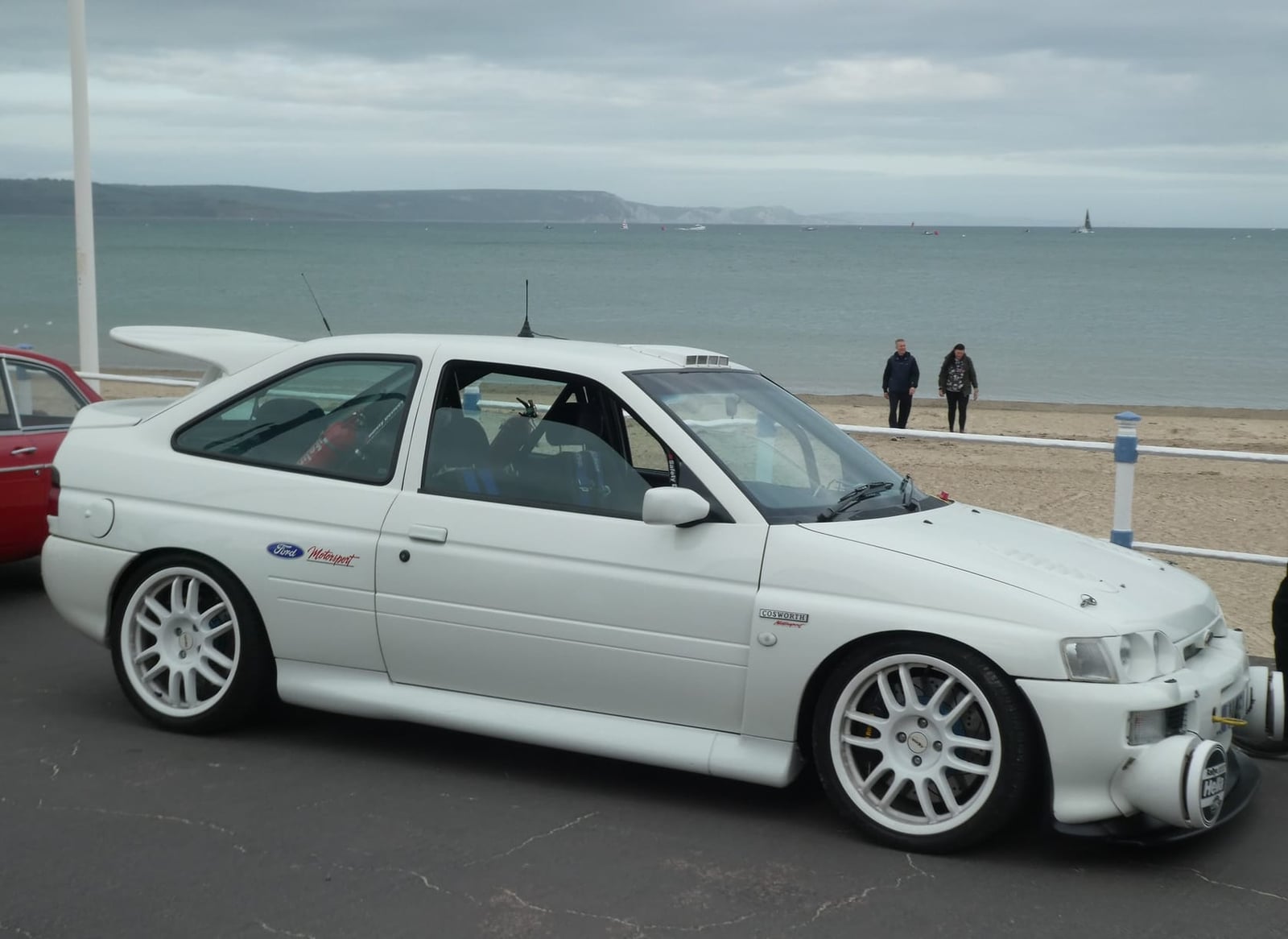 A white Ford Escort RS Cosworth in side profile at a seafront, sea behind