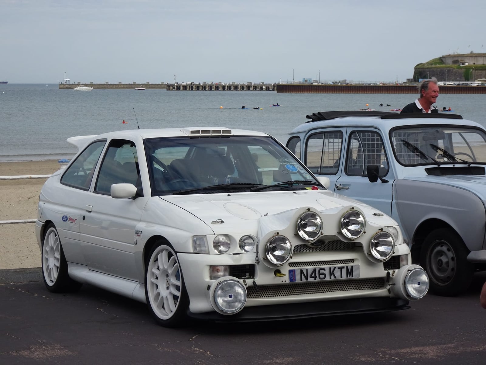 A white Ford Escort RS Cosworth in rally specification with a four-lamp pod, parked at a seafront