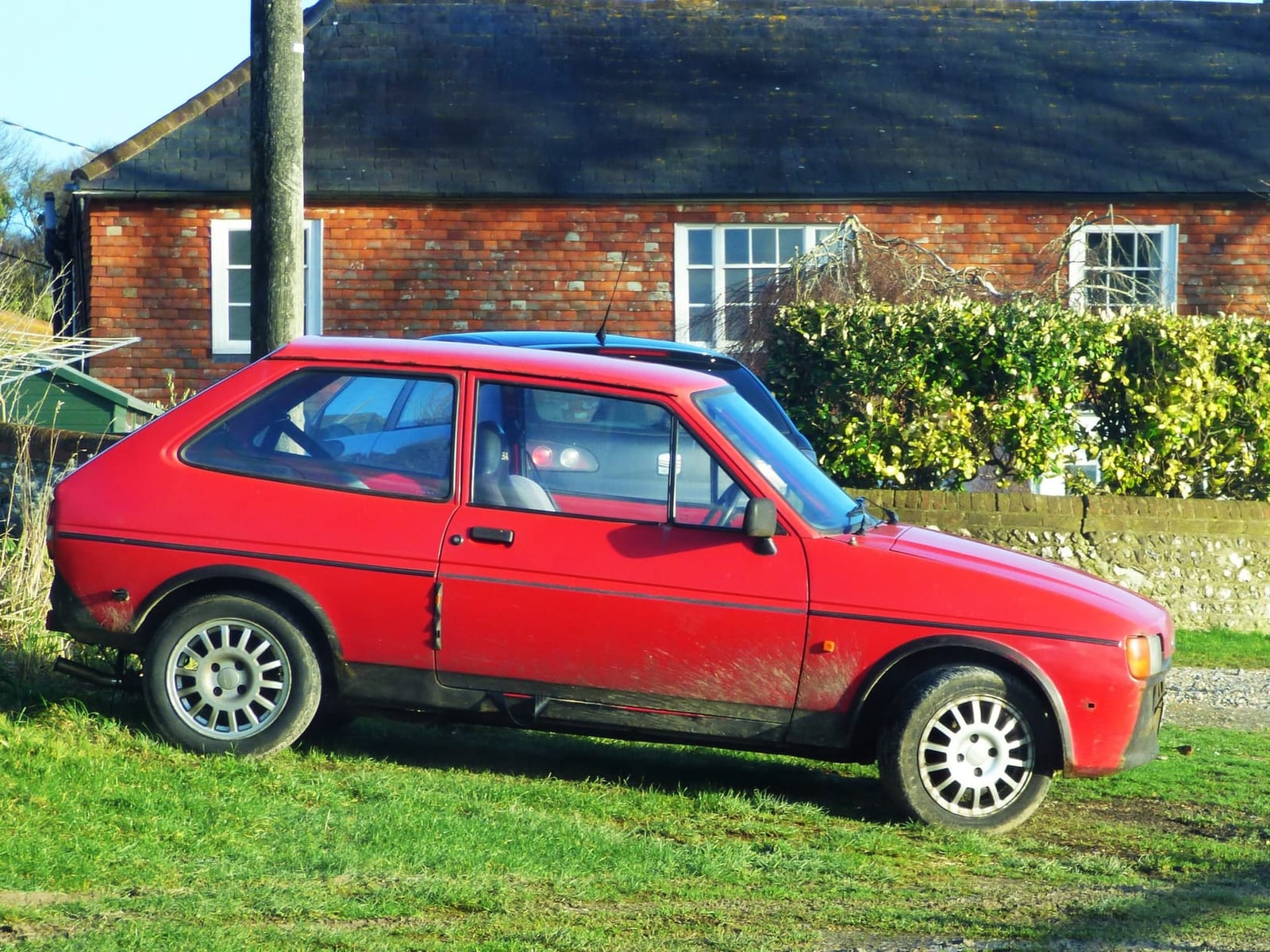 A red Ford Fiesta Mk1 parked on grass in front of a brick building, front three-quarter view