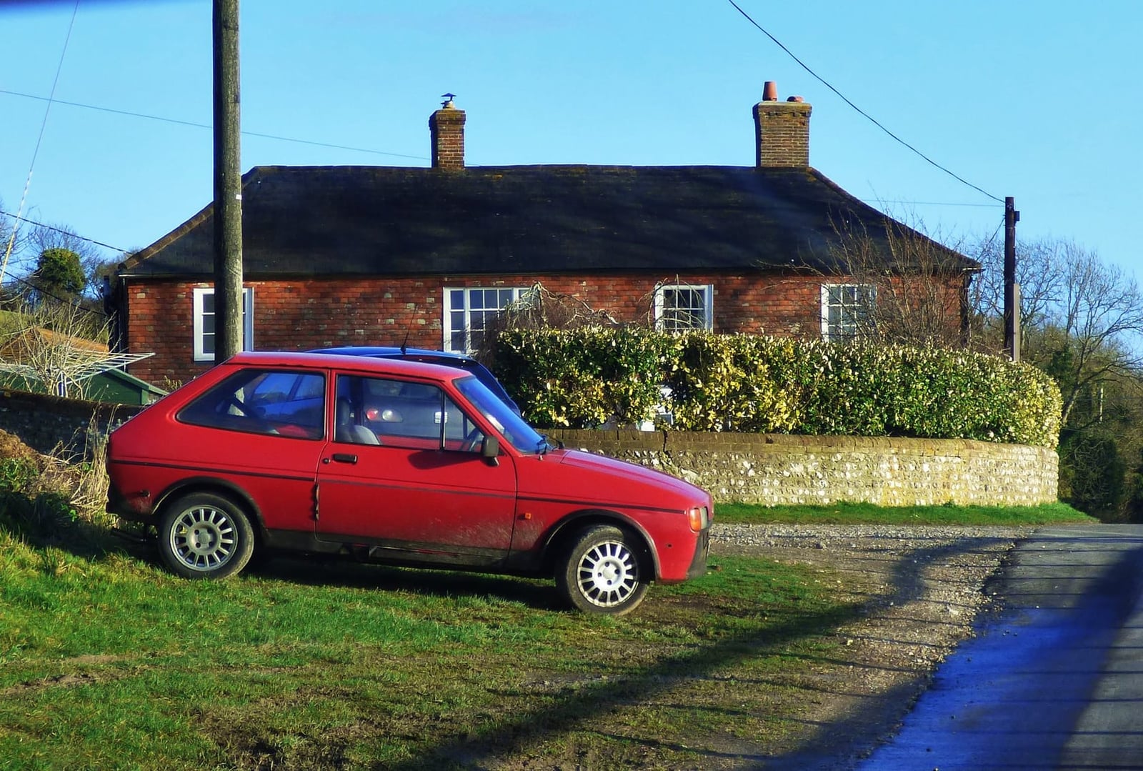 A red Ford Fiesta Mk1 parked on grass beside a hedge, side view