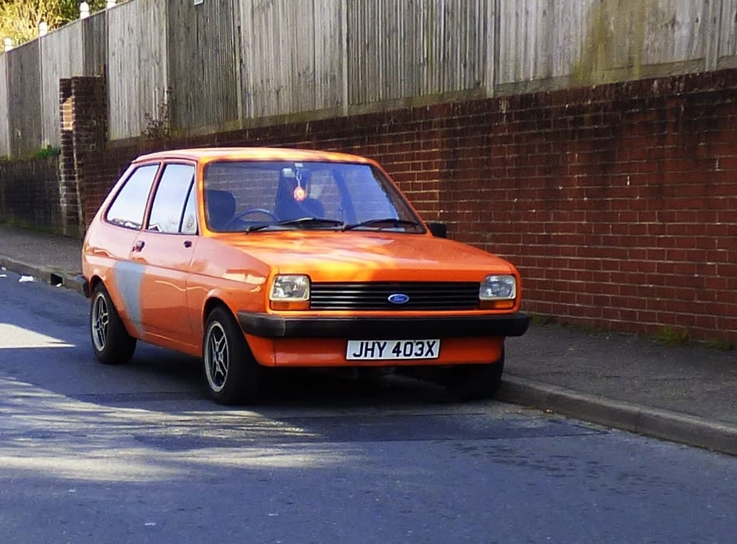 An orange Mk1 Ford Fiesta parked by a brick wall, front three-quarter view, UK registration JHY 403X