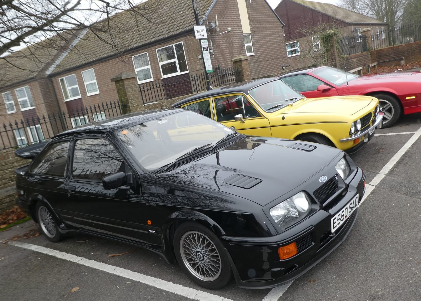 A black Ford Sierra RS500 Cosworth with a UK registration in a British car park, an orange classic and a red coupe behind