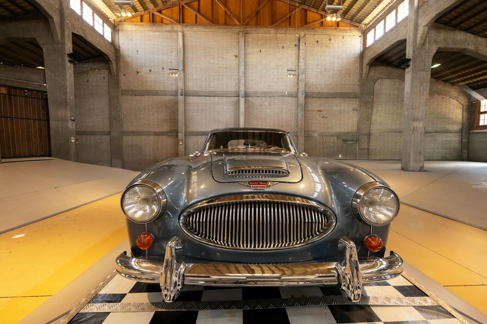 A blue Austin-Healey 3000 parked head-on, alone, in a large concrete storage hall with a timber roof