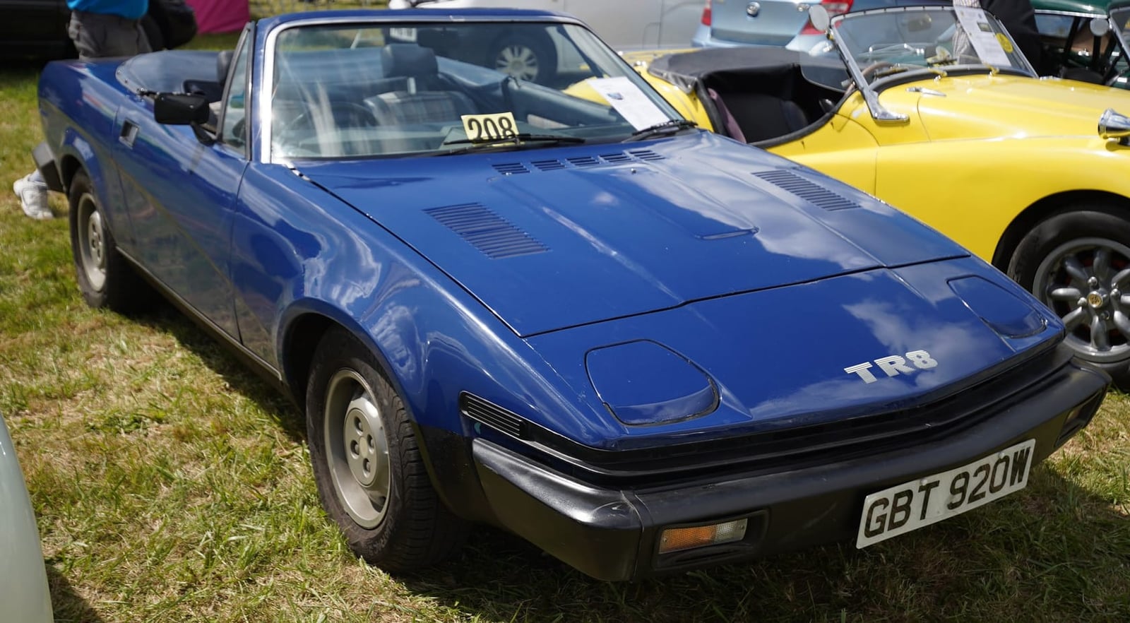 A blue Triumph TR8 convertible with a UK registration on grass, other classics parked behind