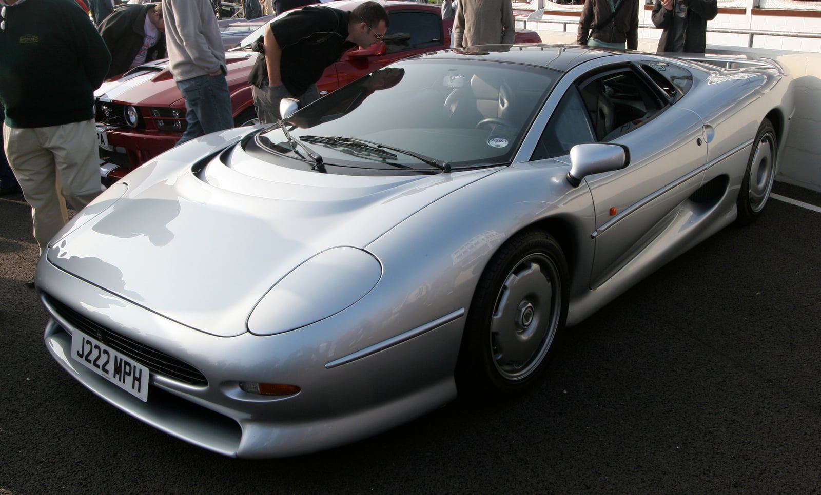 A silver Jaguar XJ220 supercar with onlookers leaning in to look, at a Goodwood Breakfast Club meet