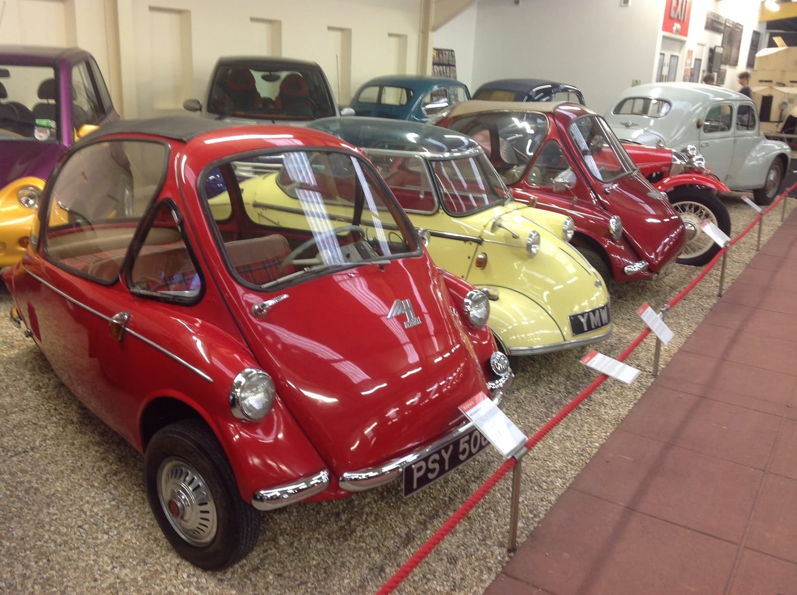 A red Heinkel bubble car leading a museum line of microcars including a yellow Messerschmitt and a Trojan