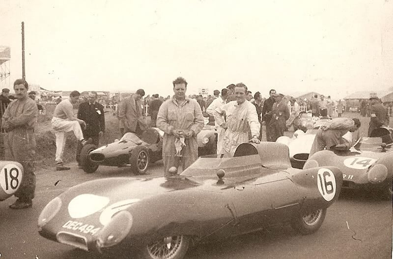 A period photograph of mechanics and drivers, including Graham Hill, around Lotus Eleven racing cars in a paddock
