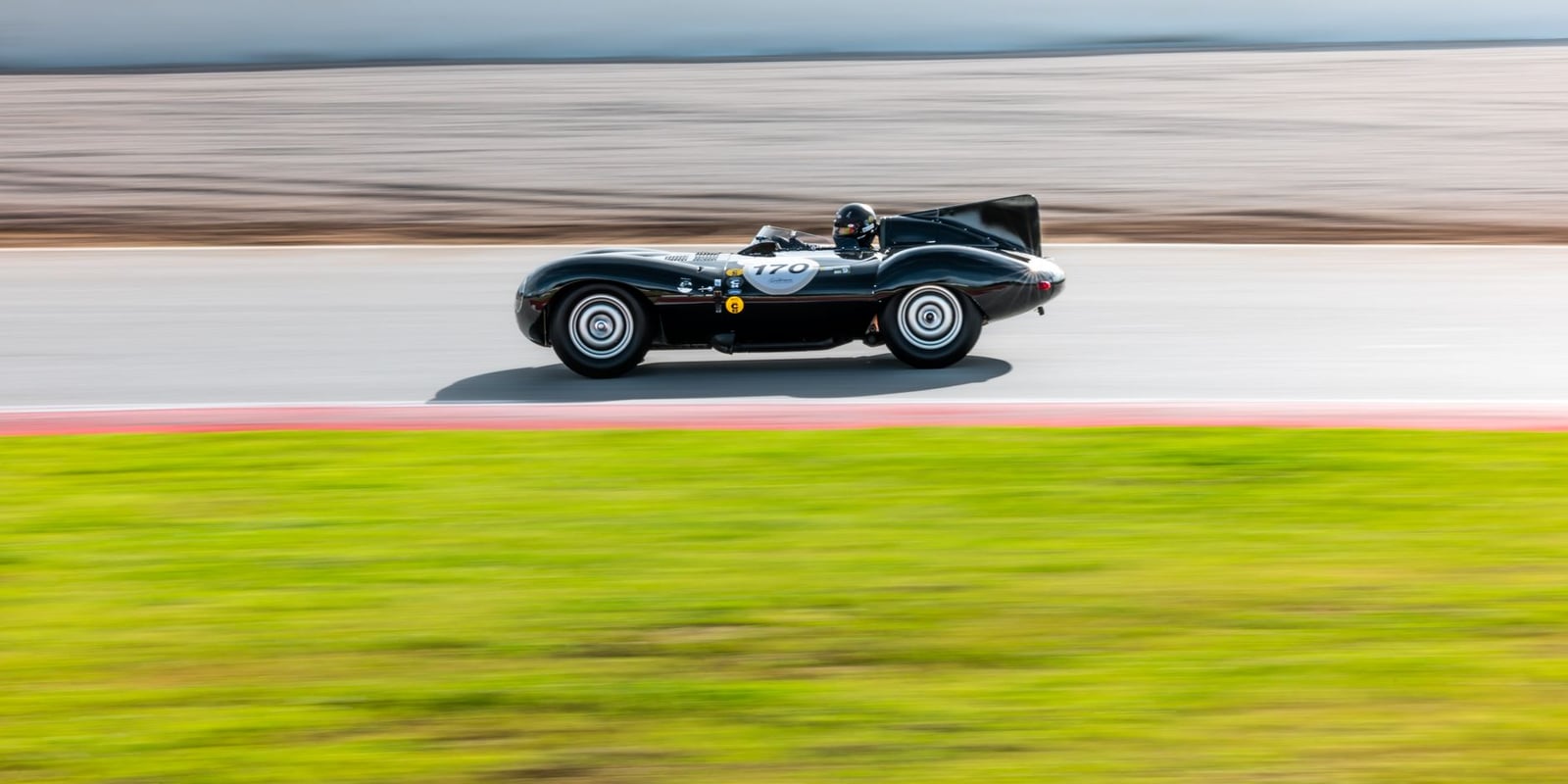 A black Jaguar D-Type historic racing car at speed in side profile on a circuit, the background blurred by panning