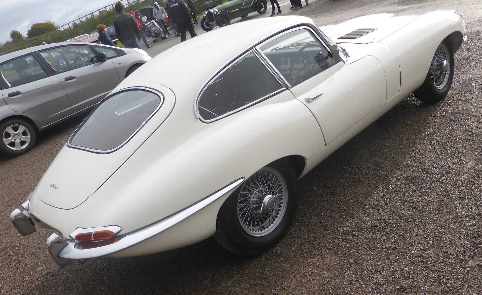 A white 1962 Jaguar E-Type 3.8 fixed-head coupé, rear three-quarter view, parked among other cars at a show