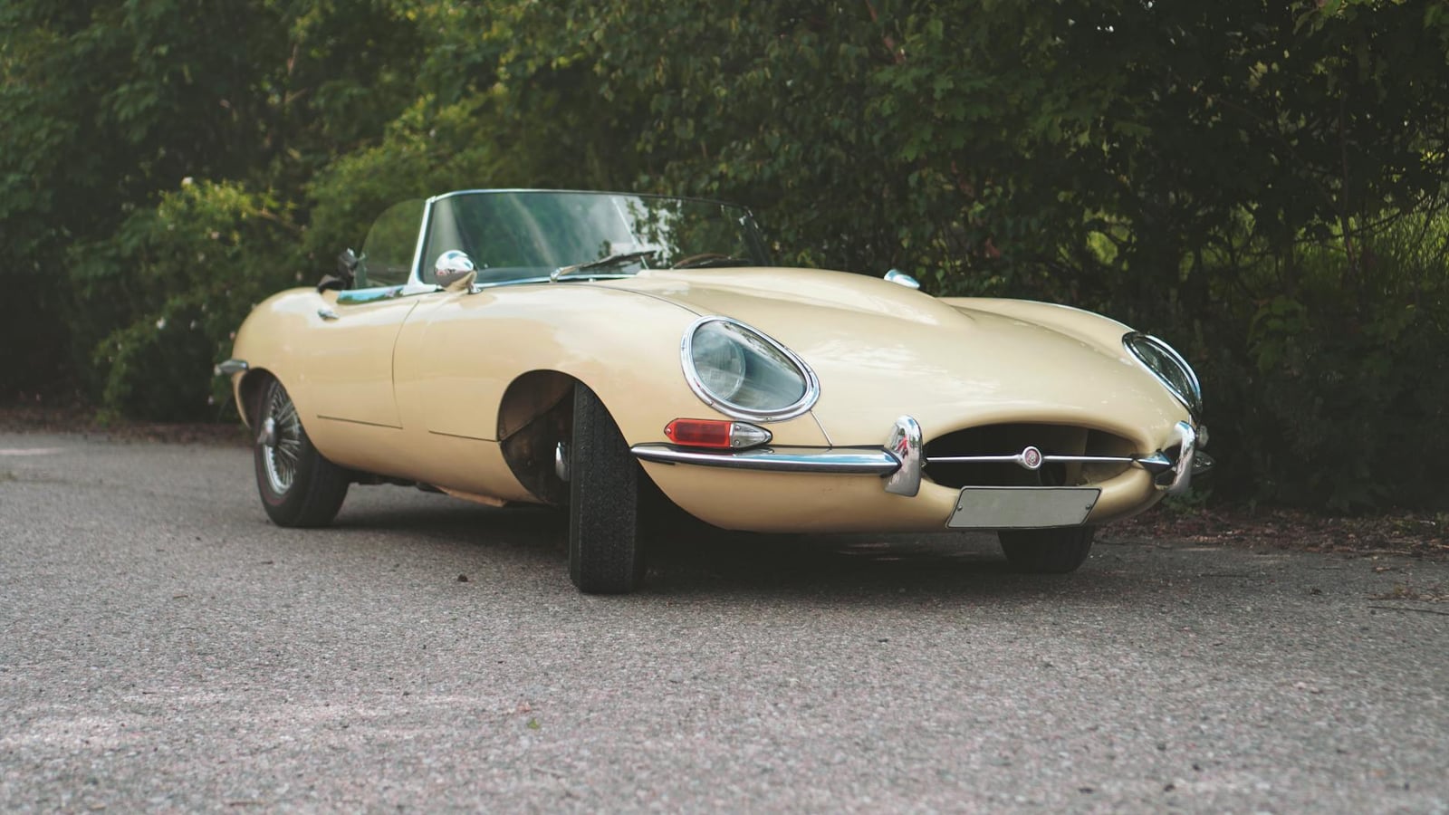 A beige Jaguar E-Type roadster parked on a quiet road, front three-quarter view