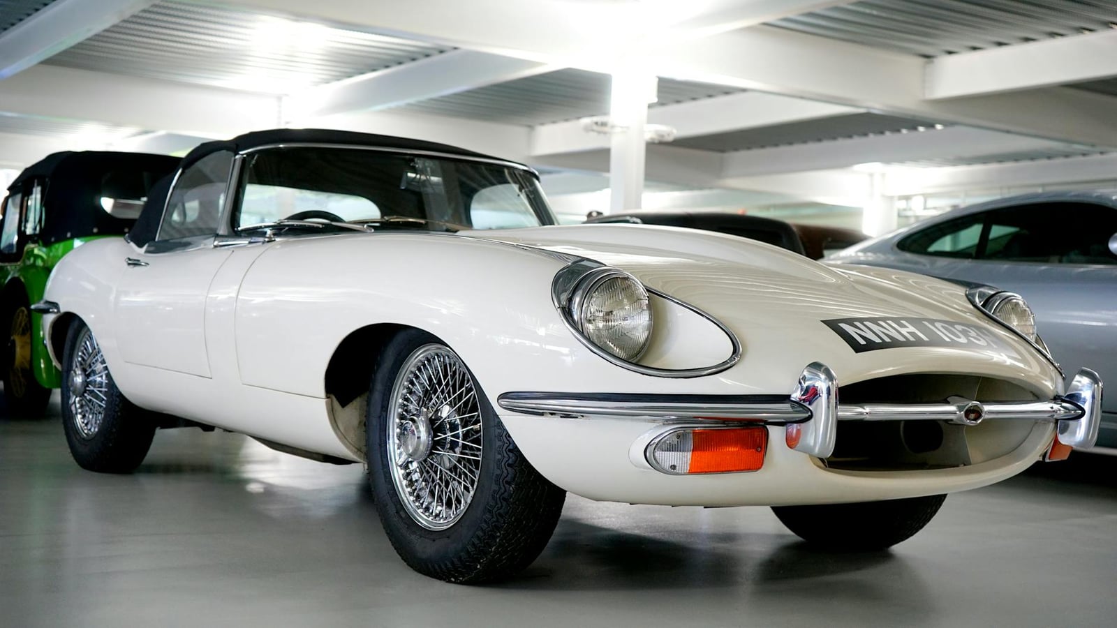 A white Jaguar E-Type roadster with open headlamps and slim chrome bumpers displayed indoors