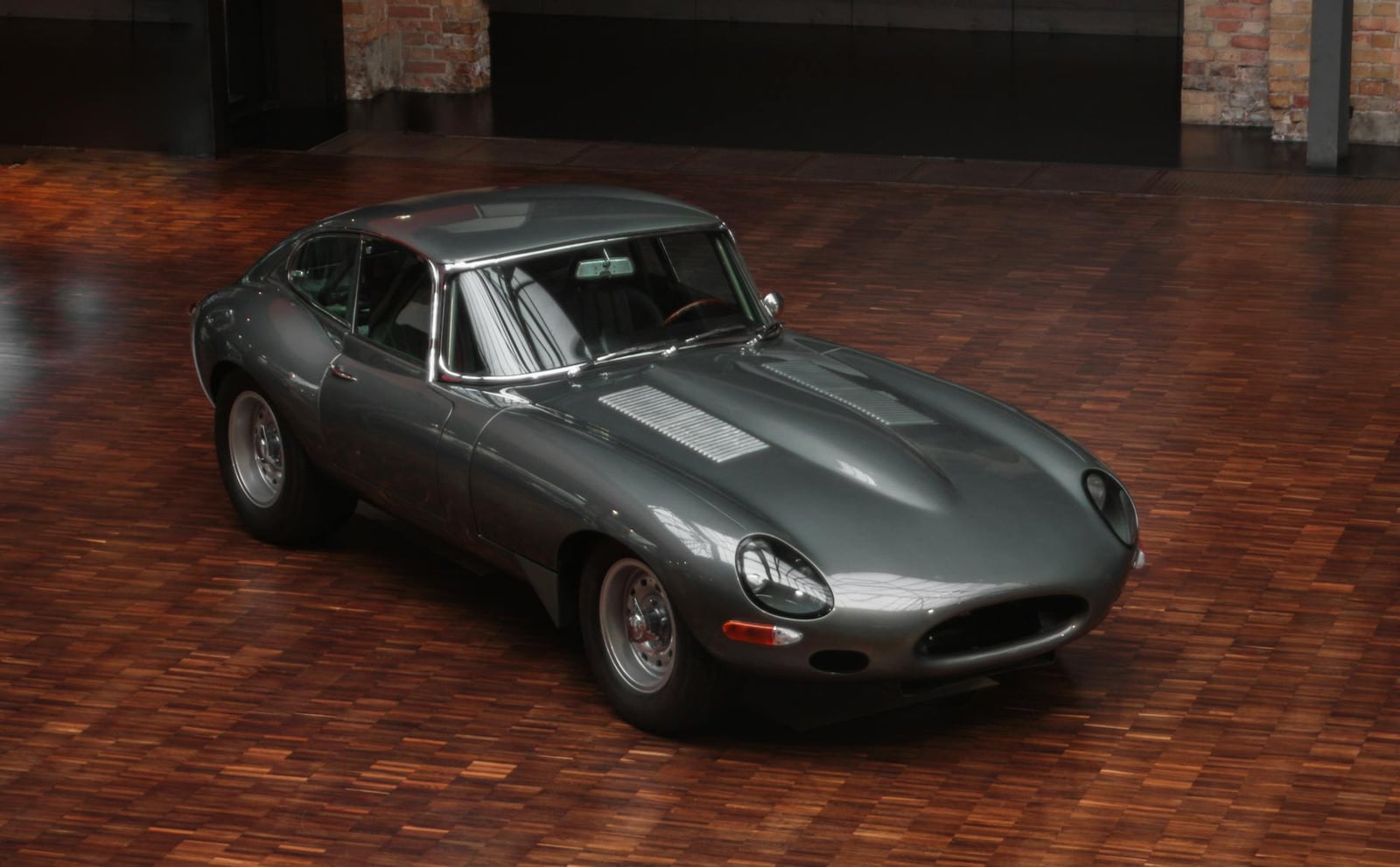 A grey Jaguar E-Type Series 1 fixed-head coupe with covered headlamps displayed indoors