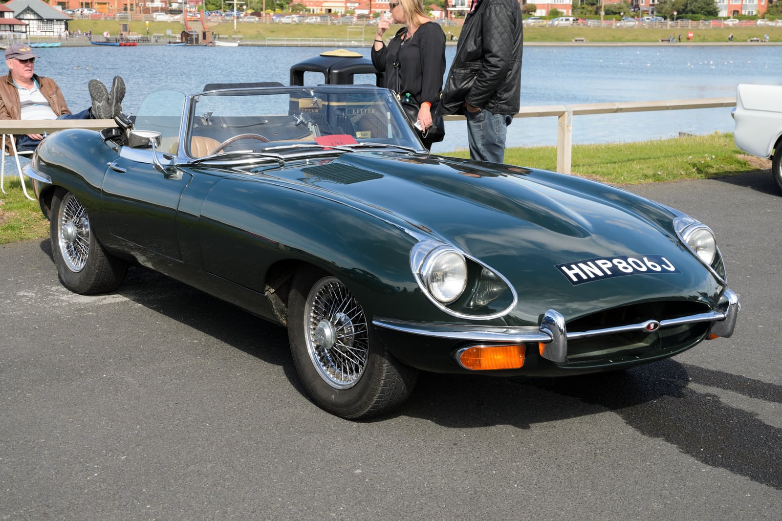 A dark green Jaguar E-Type Series 2 roadster with open headlamps parked beside water