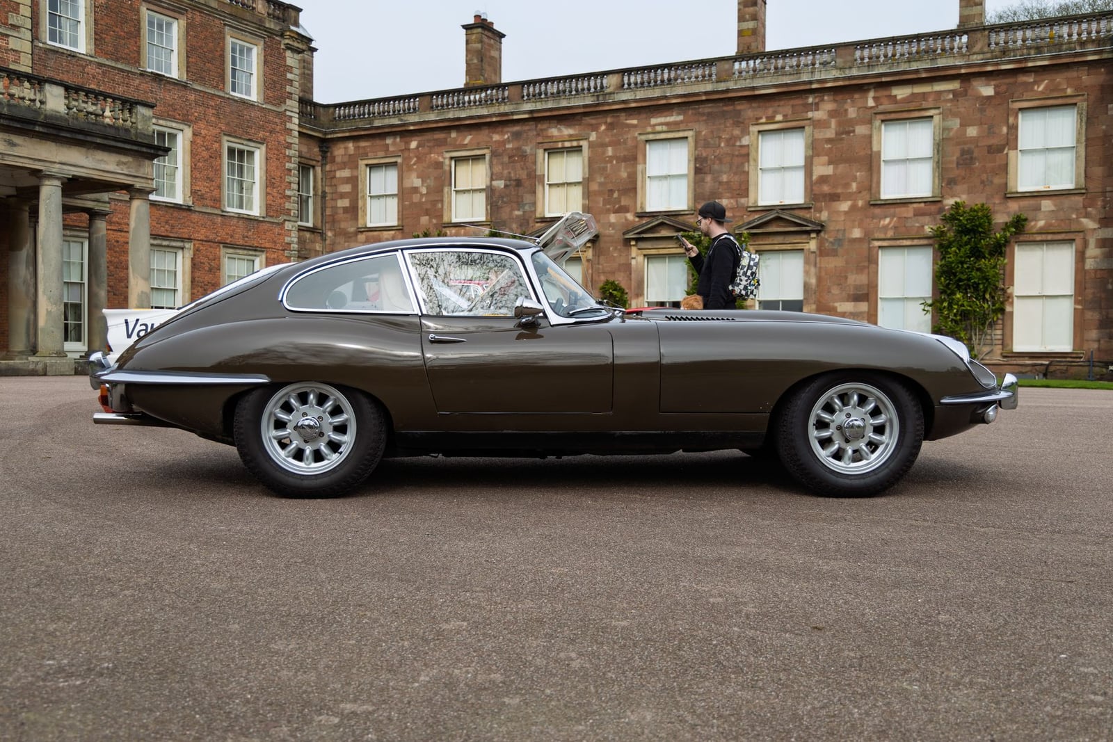 A gunmetal grey Jaguar E-Type fixed-head coupe in front of a country house, side view
