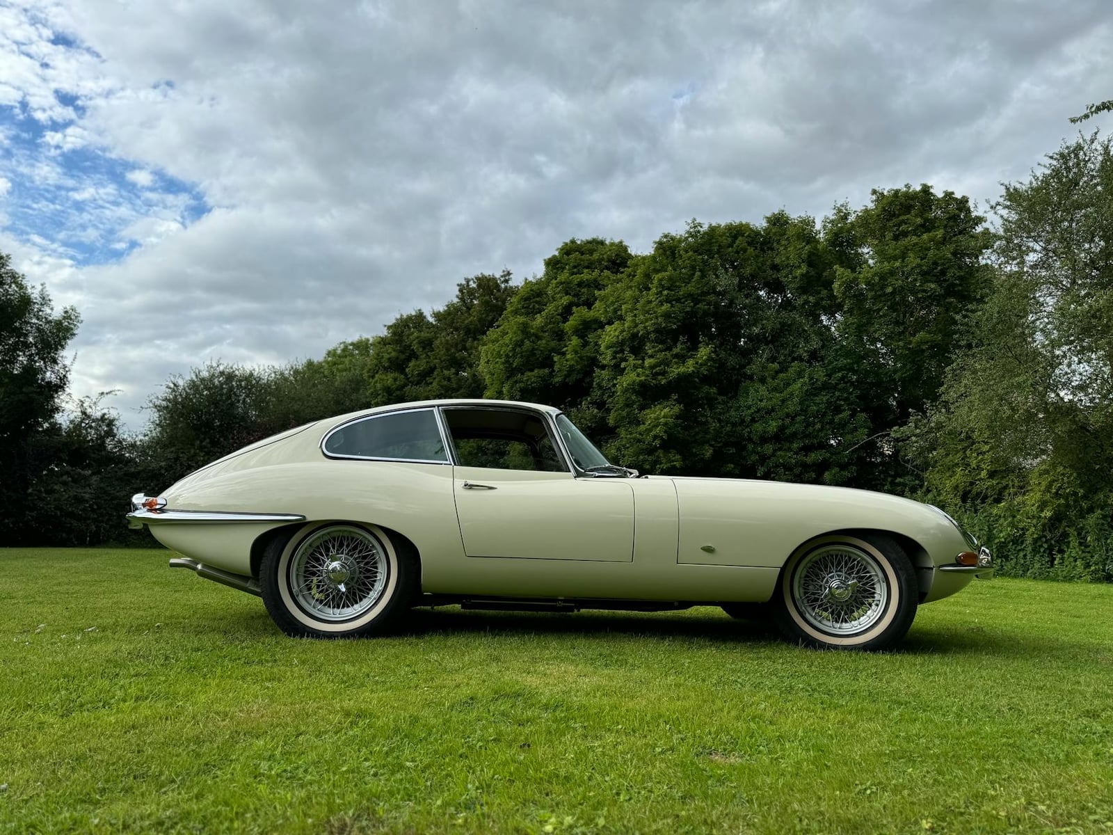 A cream Jaguar E-Type Series 1 fixed-head coupe with covered headlamps parked on a lawn, side view