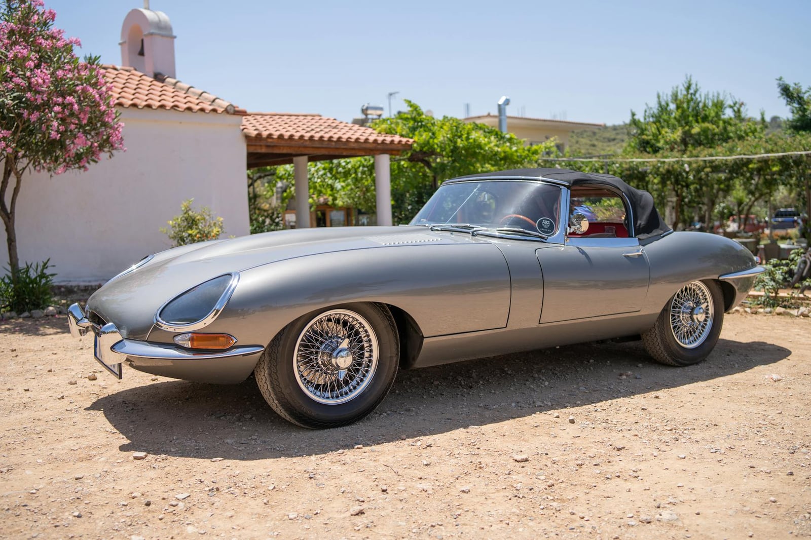 A silver Jaguar E-Type Series 1 roadster with covered headlamps and wire wheels parked in a sunny yard, side view