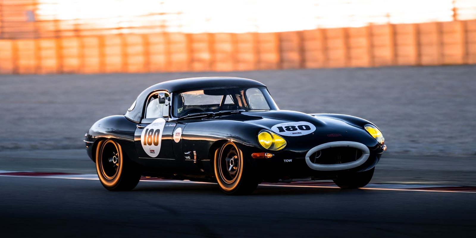 A dark blue Jaguar E-Type lightweight racing car, number 100 on gold wheels, cornering on a circuit at dusk