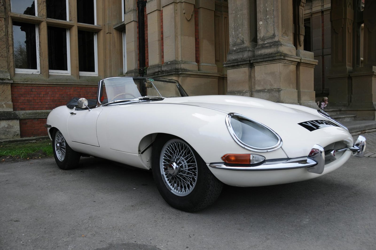 A white Jaguar E-Type Series 1 roadster with wire wheels parked beside a historic stone building, front three-quarter view