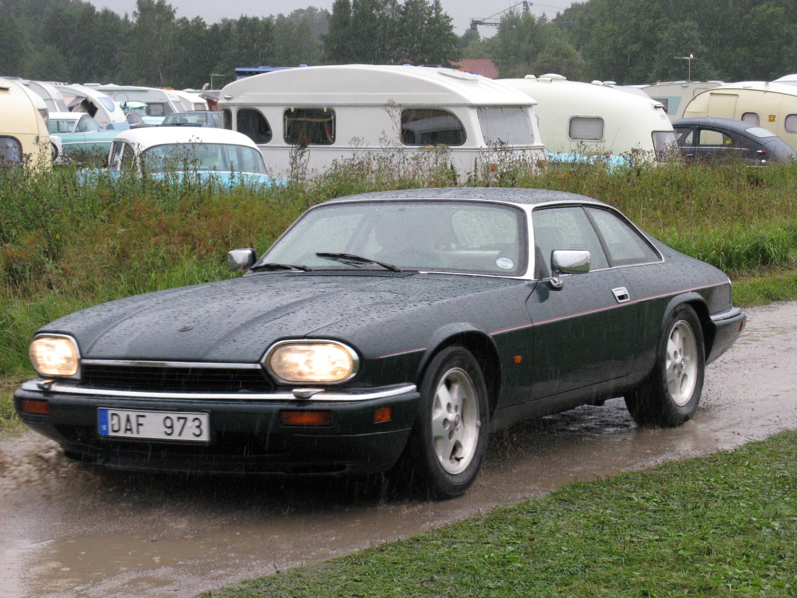 A dark Jaguar XJS fixed-head coupe parked on a wet road, front three-quarter view