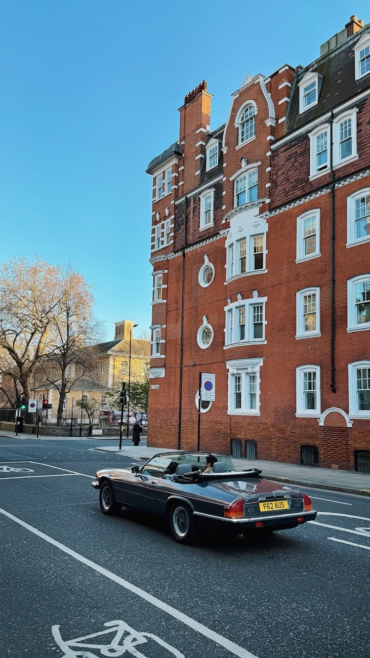 A black Jaguar XJS convertible driving past a red-brick mansion block on a London street