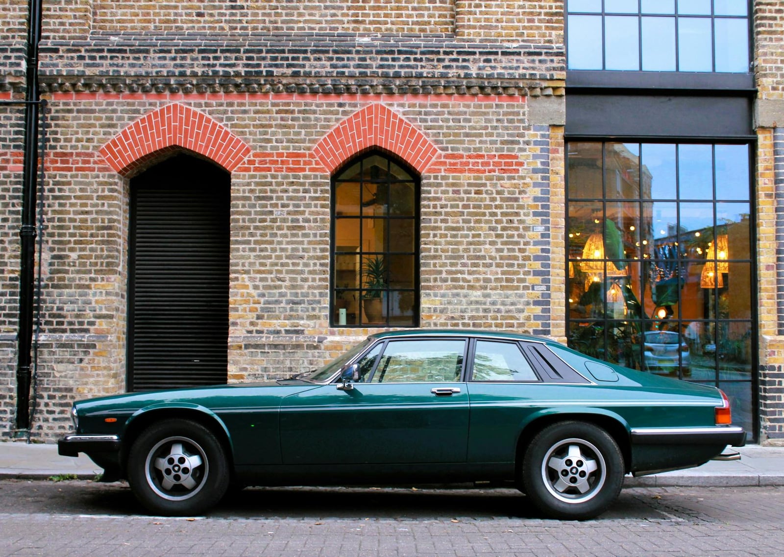 A dark green Jaguar XJS coupe in side profile, parked by a brick building