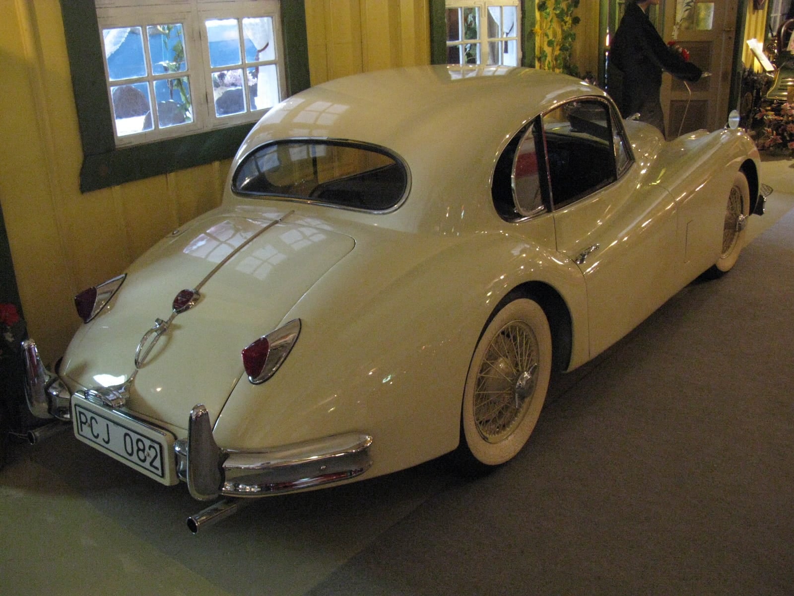 A cream 1956 Jaguar XK140 fixed-head coupe indoors, rear three-quarter view, UK registration FCJ 082