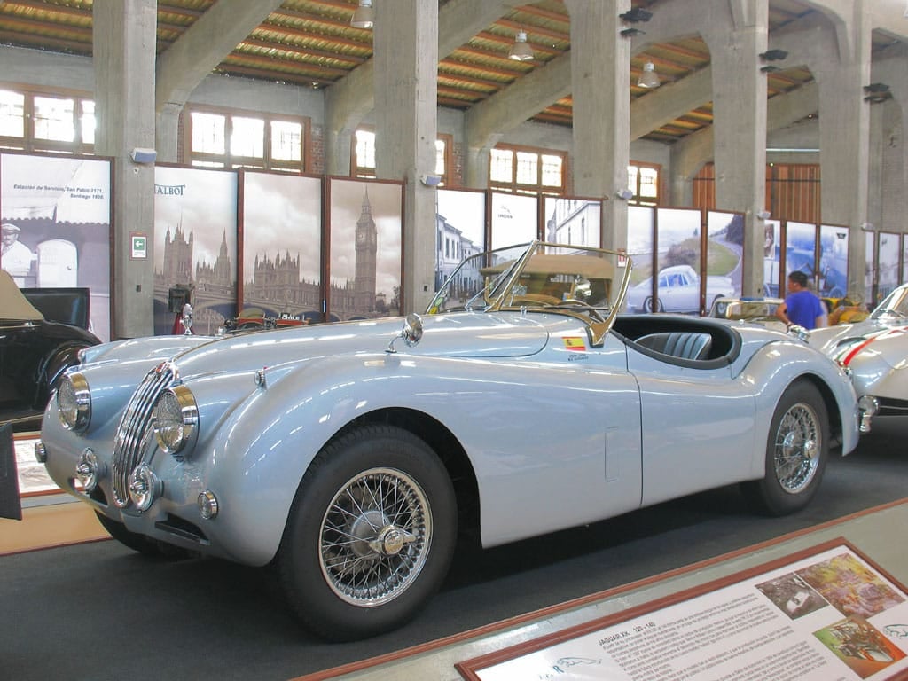 A pale blue 1955 Jaguar XK140 roadster displayed in a museum hall