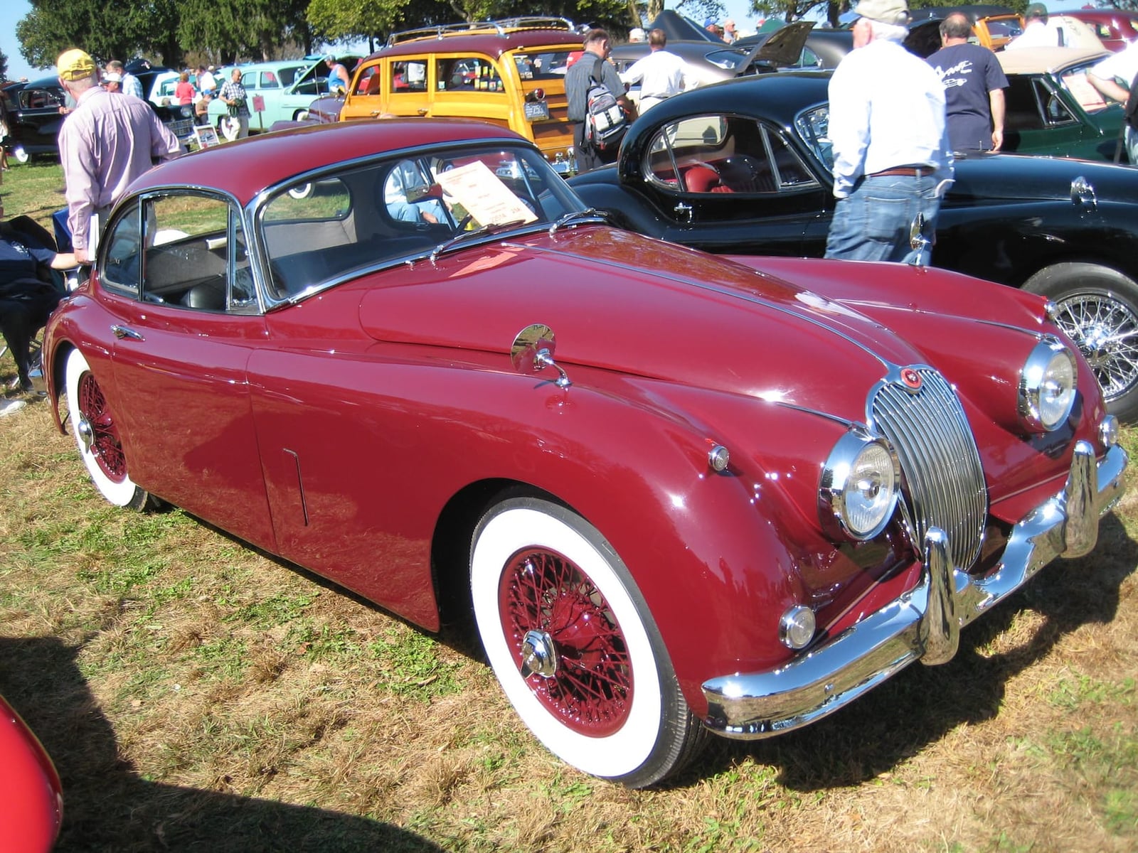 A maroon Jaguar XK150 fixed-head coupe with whitewall tyres and wire wheels on grass at a classic car show