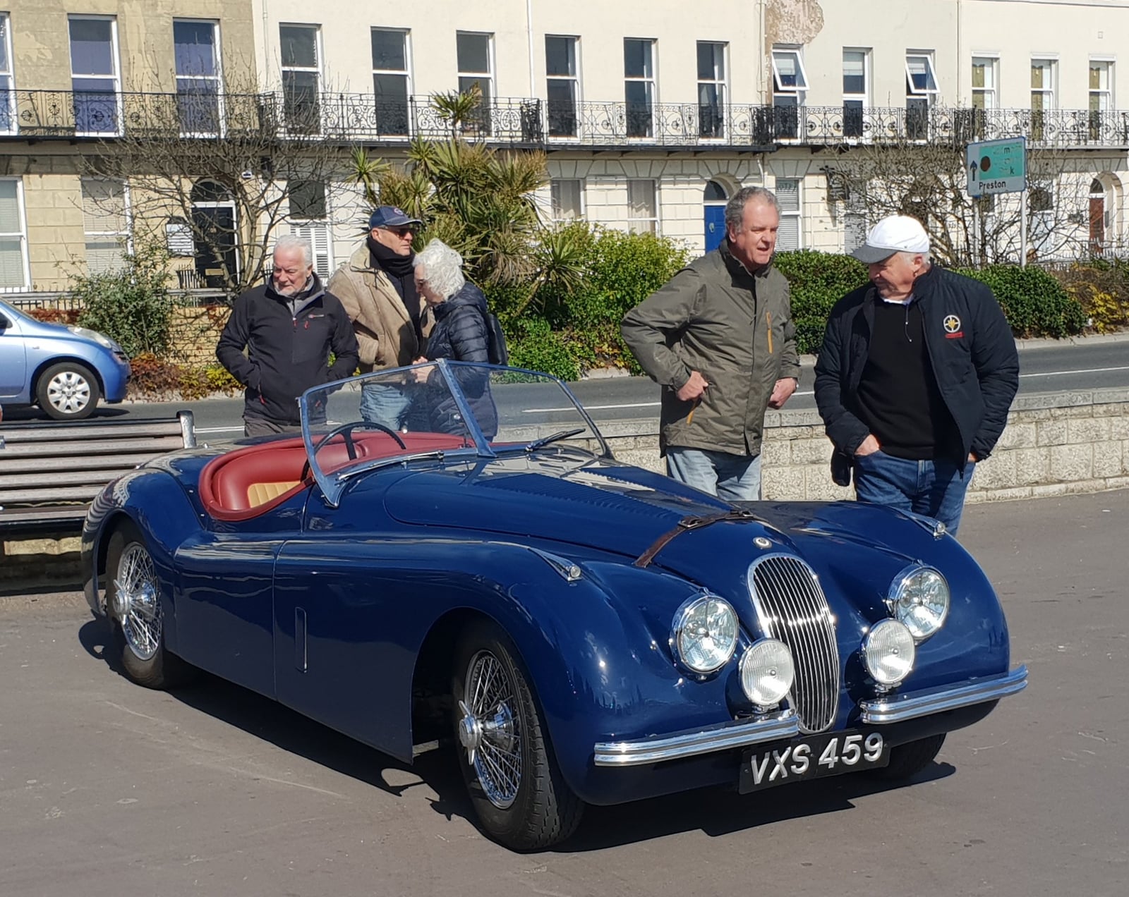 A dark blue Jaguar XK120 open roadster with red interior, front three-quarter view on a seafront, UK registration VXS 459