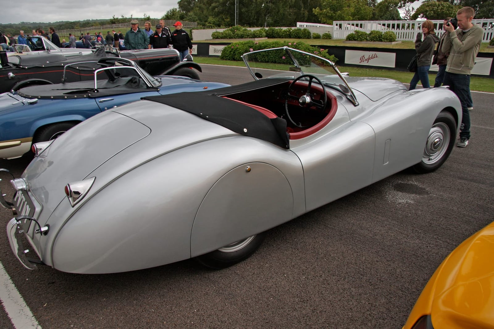 A silver Jaguar XK120 roadster seen from the rear three-quarter at Goodwood, its tonneau cover fitted over the passenger side