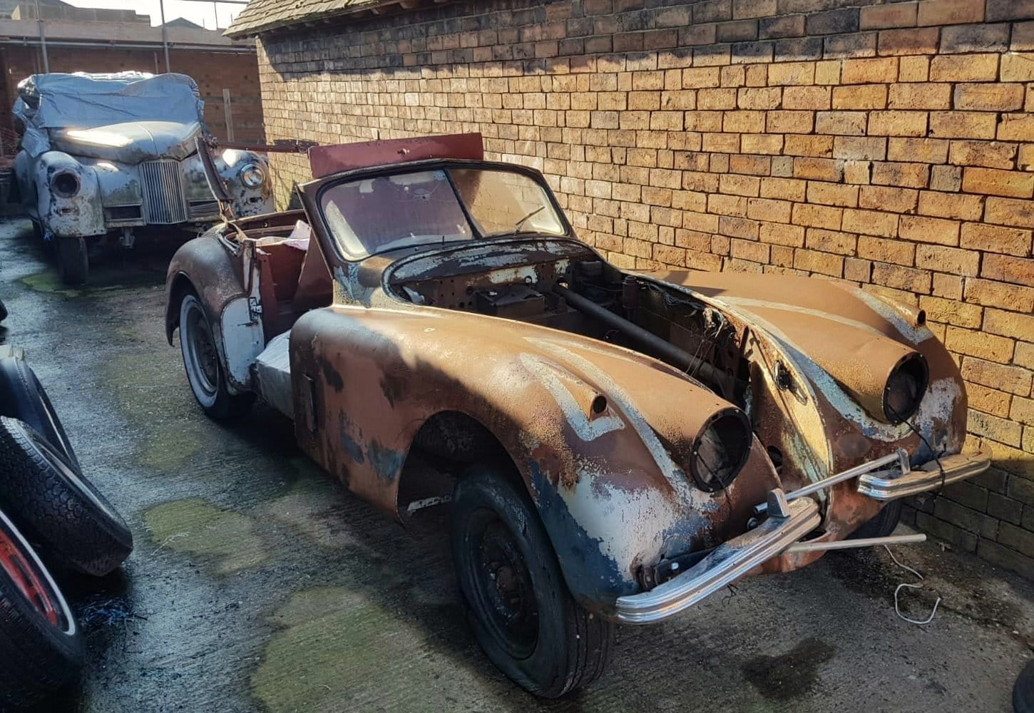 A stripped and unrestored Jaguar XK140 roadster project in bare patinated metal beside a brick wall