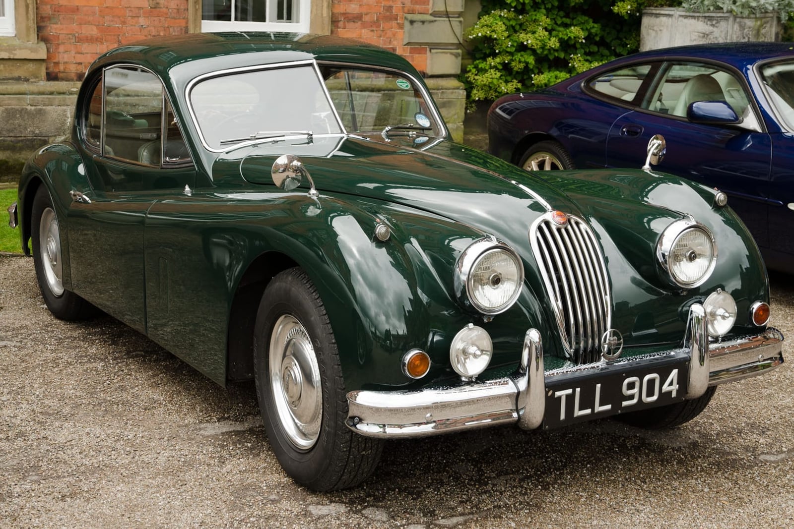 A dark green 1956 Jaguar XK140 fixed-head coupé with a UK registration parked on gravel