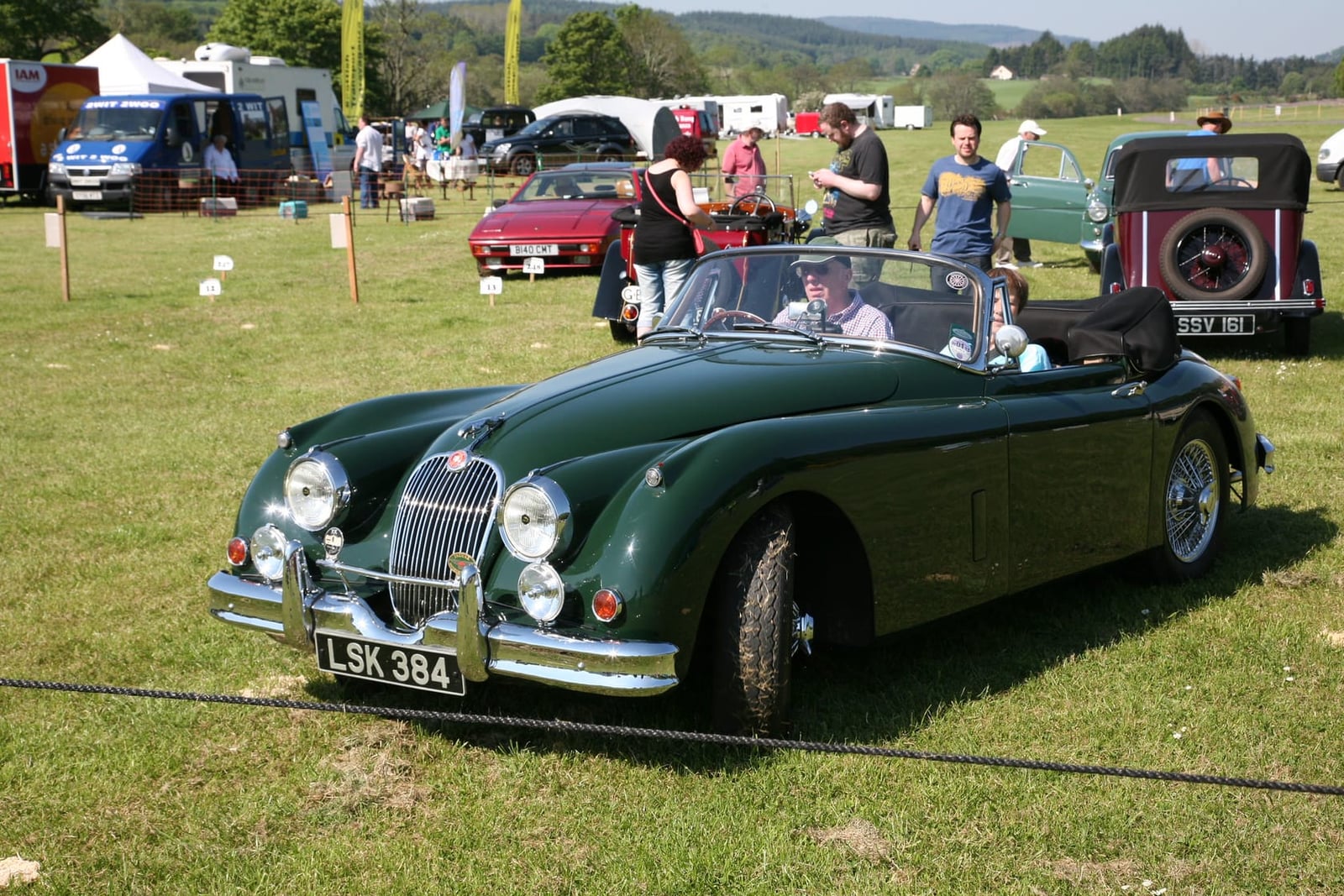 A dark green Jaguar XK150 roadster on grass at a classic car show with the hood down, UK registration LSK 384