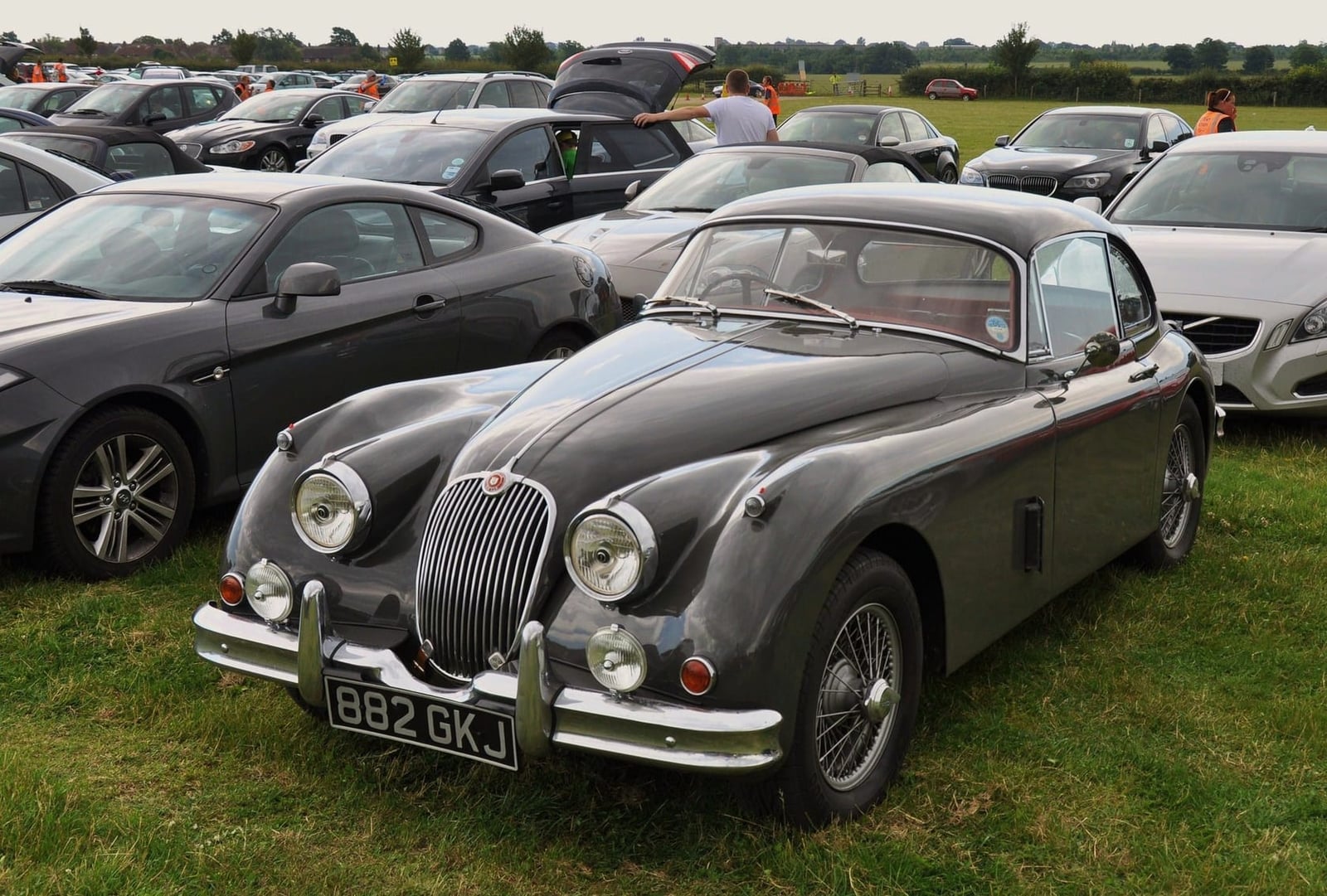 A grey Jaguar XK150 fixed head coupe parked among modern cars, front three-quarter view, UK registration 882 GKJ