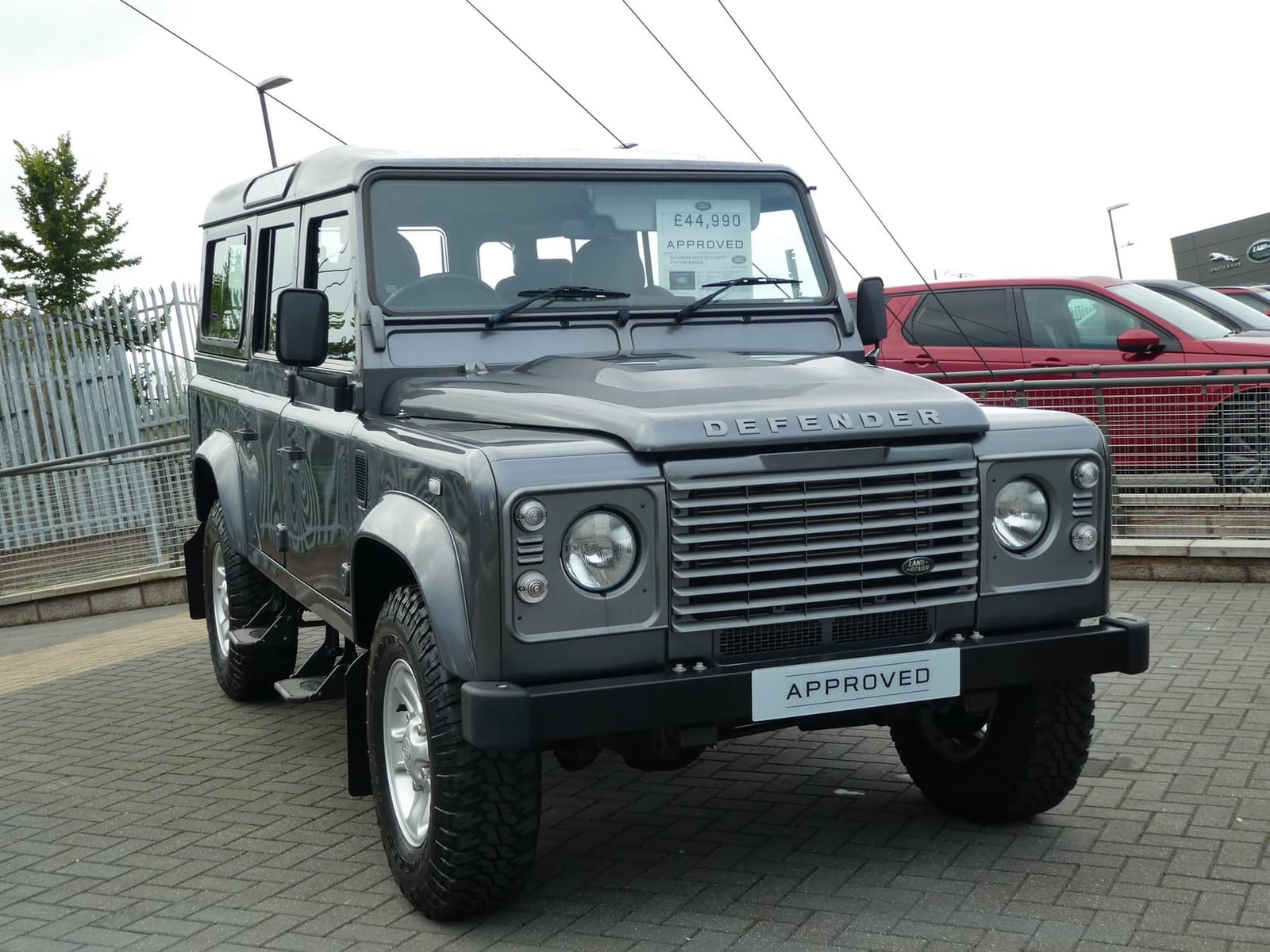 A grey 2015 Land Rover Defender 110 County station wagon on a dealer forecourt with an approved-used plate and a price in the windscreen