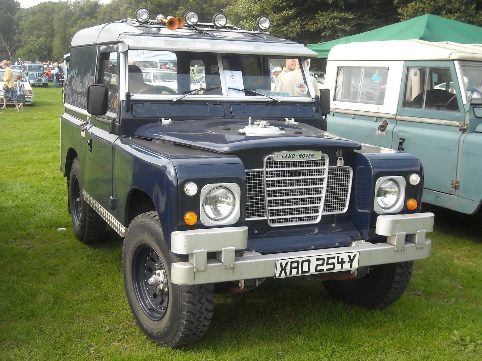A dark blue classic Land Rover with roof-mounted spotlights at an outdoor show, another early Land Rover parked beside it