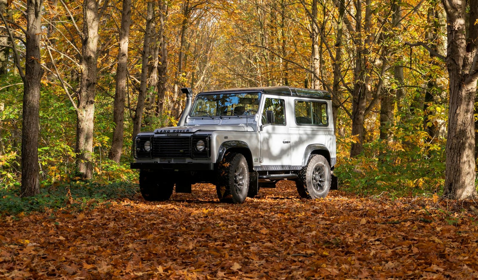A silver Land Rover Defender 110 parked on a leaf-strewn track in an autumn forest