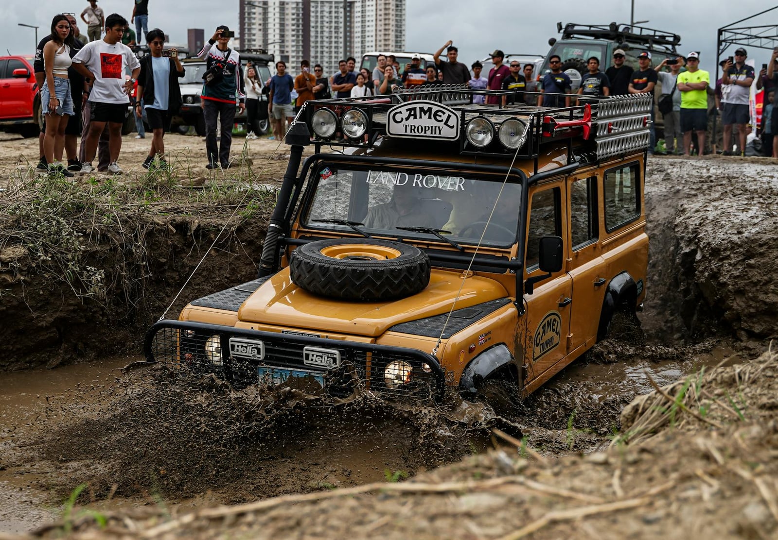 A sand-coloured Camel Trophy Land Rover Defender 110 wading through deep mud