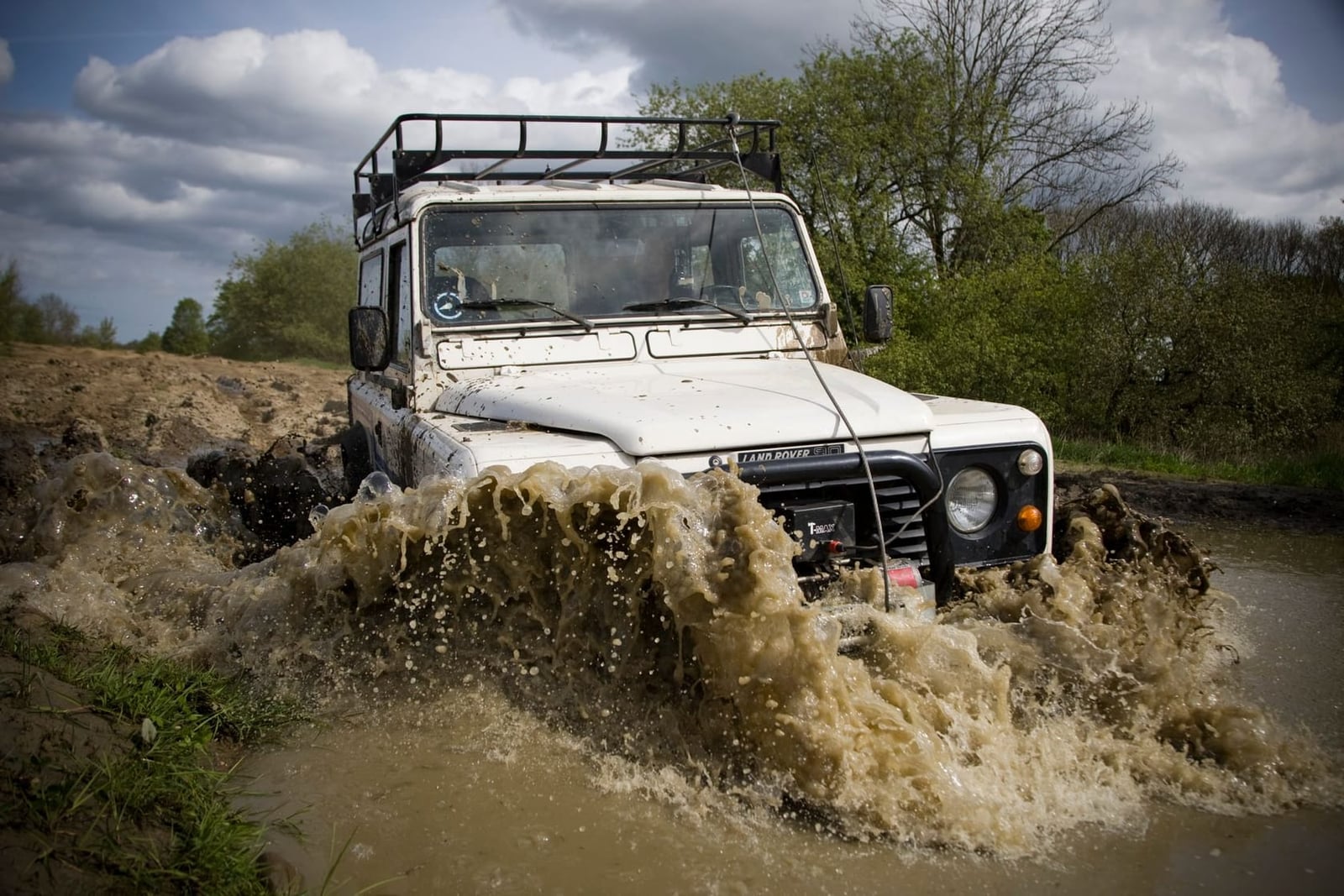 A white Land Rover Defender 110 with a roof rack wading through deep muddy water on an off-road course