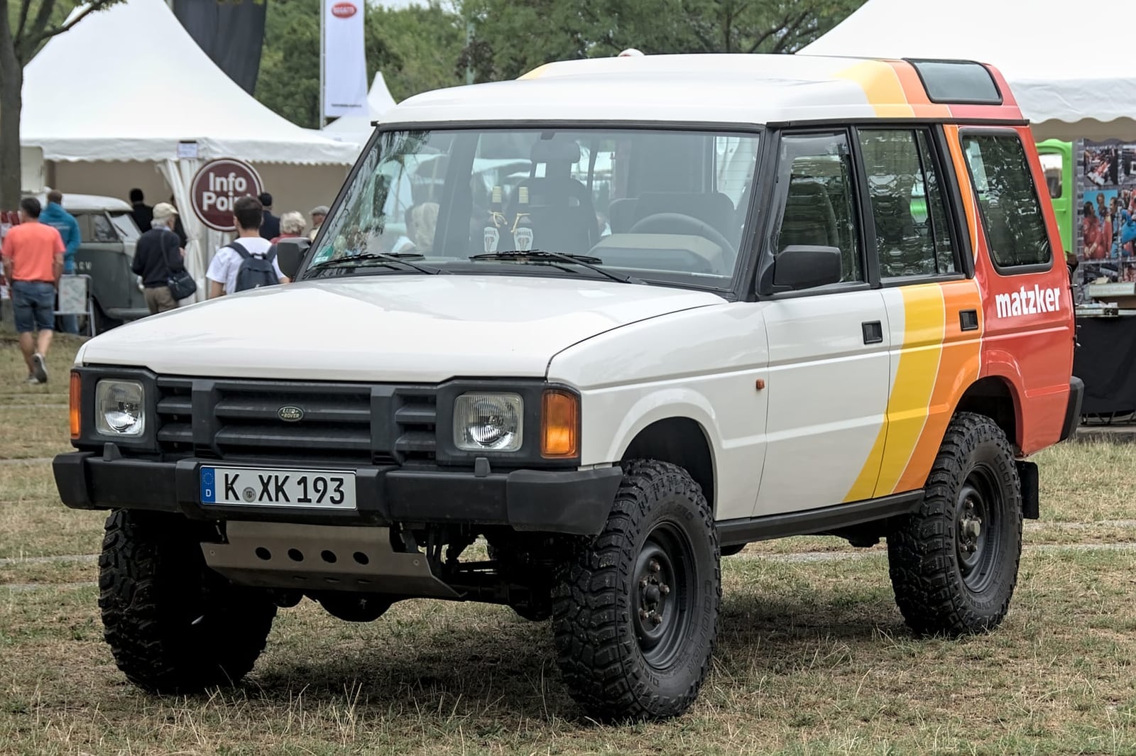A white and orange expedition-liveried Land Rover Discovery 1 with roof rack and off-road tyres on a show field