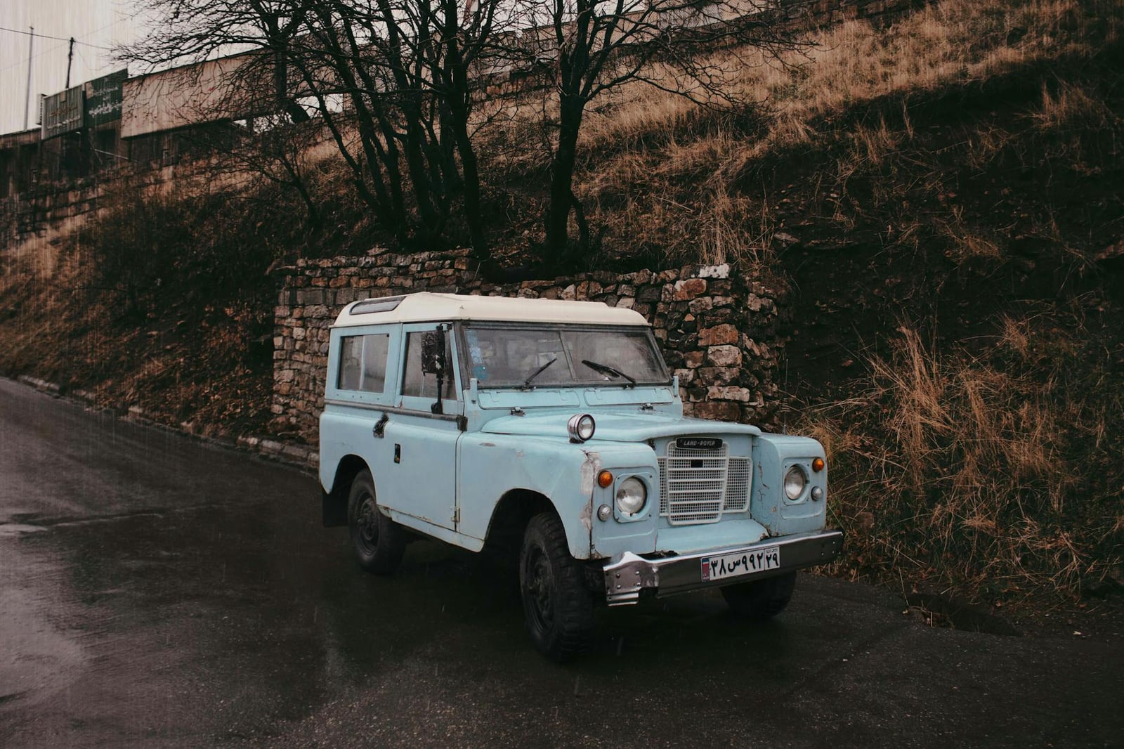 A light blue Land Rover Series III parked on a wet roadside below a hill