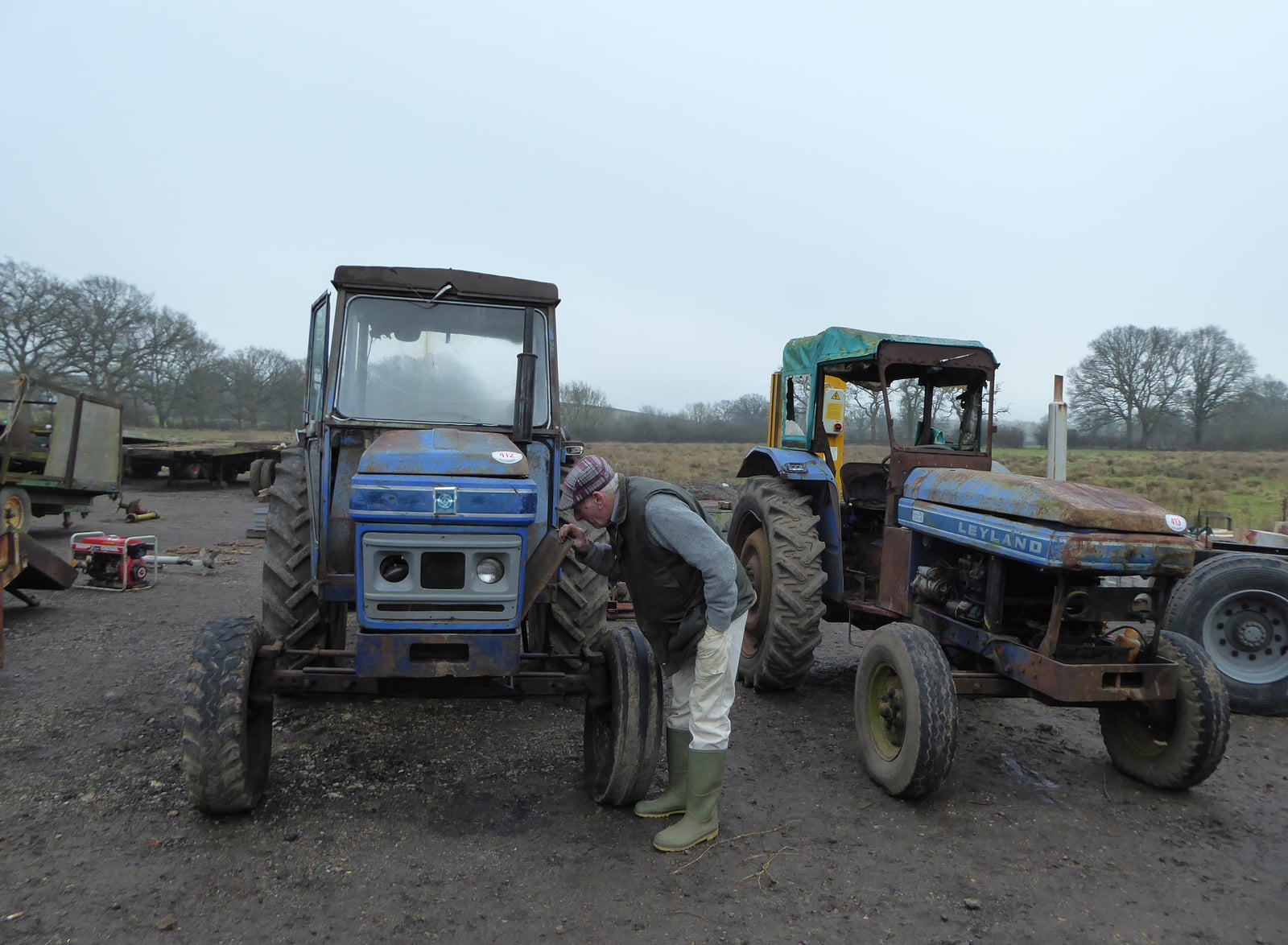 A man in a flat cap inspecting the front of a blue Leyland tractor at a farm sale, a second Leyland alongside on a misty day