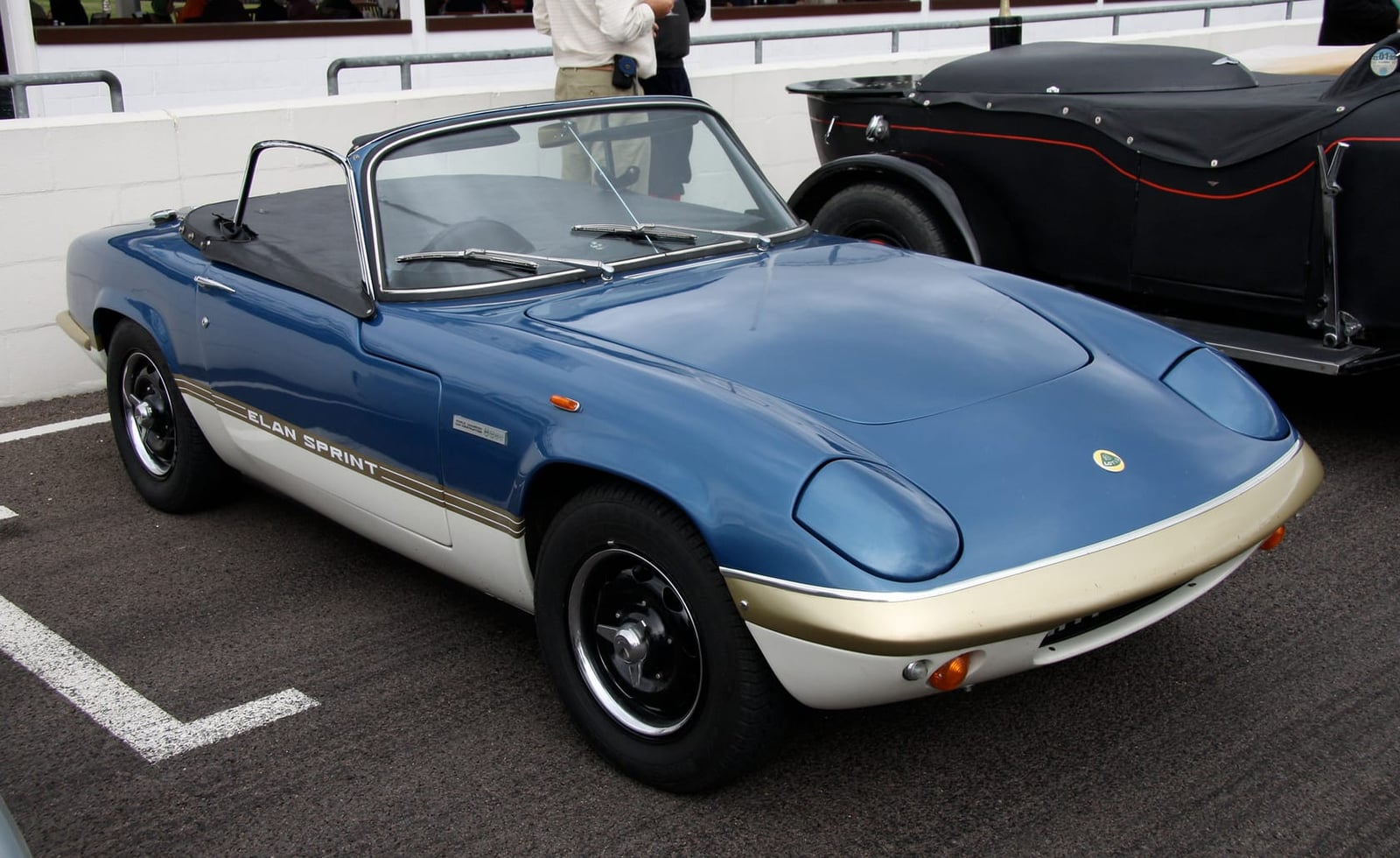 A blue Lotus Elan roadster with gold sill stripes parked on tarmac, front three-quarter view