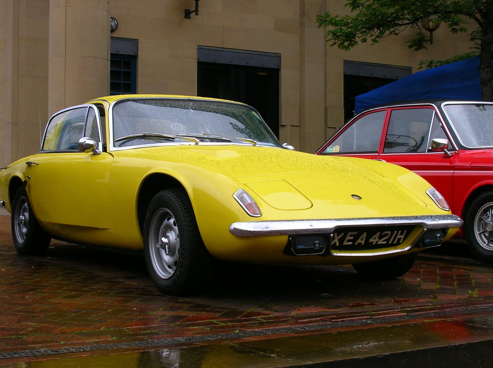 A yellow Lotus Elan +2 coupe parked outside a building, front three-quarter view, UK registration KEA 421H