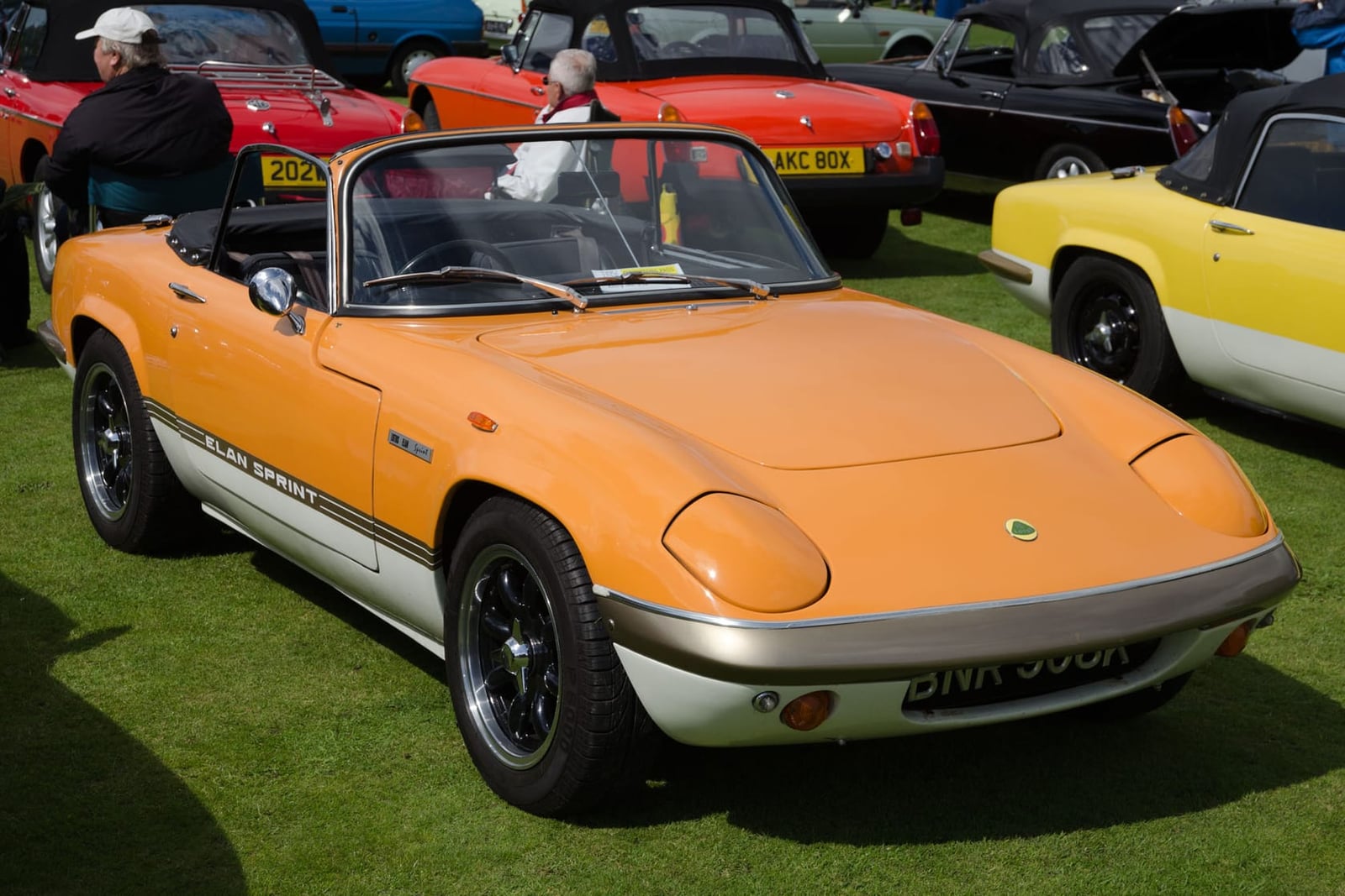 An orange and gold two-tone 1971 Lotus Elan Sprint on grass, front three-quarter view